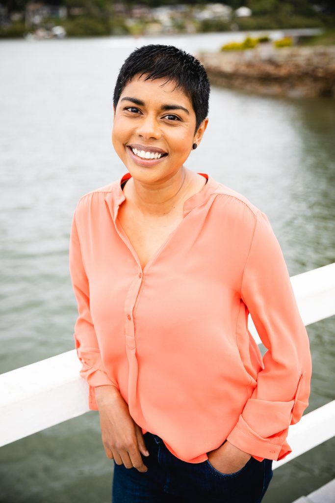 Colour publicity photo of Dinuka McKenzie smiling while leaning against a white wharf fence with the river in the background. Dinuka has short black hair, brown skin and is wearing a coral long sleeved top and blue jeans.