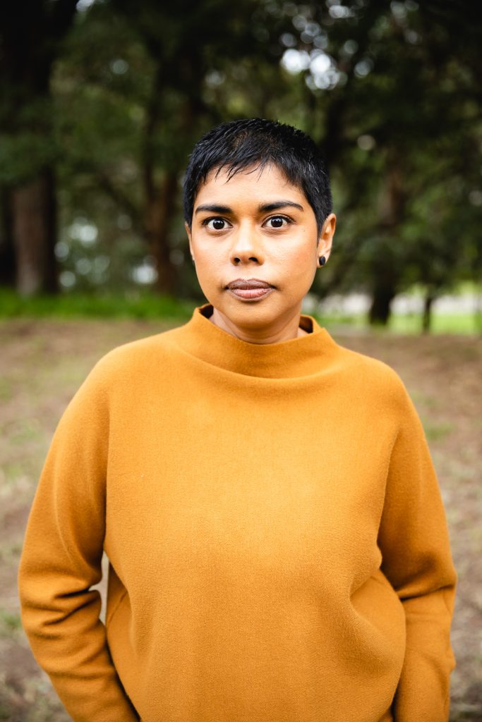 Colour publicity photo of Dinuka McKenzie from the waist up with bushland in the background. Dinuka has short black hair, brown skin and is wearing a tan coloured long sleeved top,