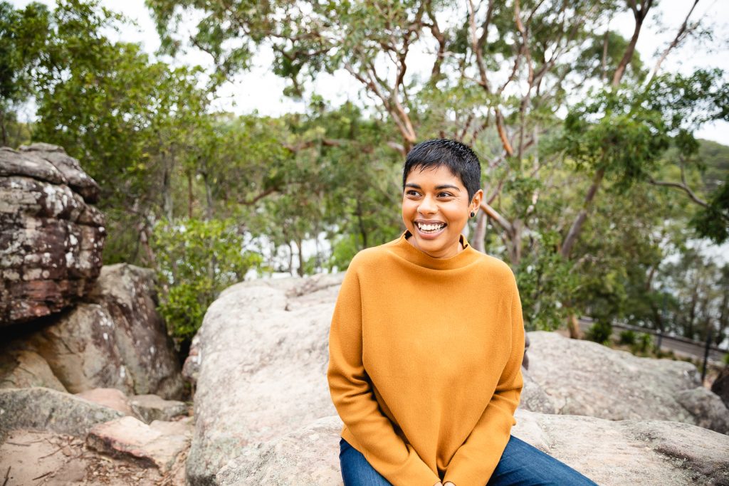 Colour publicity photo of Dinuka McKenzie laughing while sitting on rocks with bushland in the background. Dinuka has short black hair, brown skin and is wearing a tan long sleeved top and blue jeans.