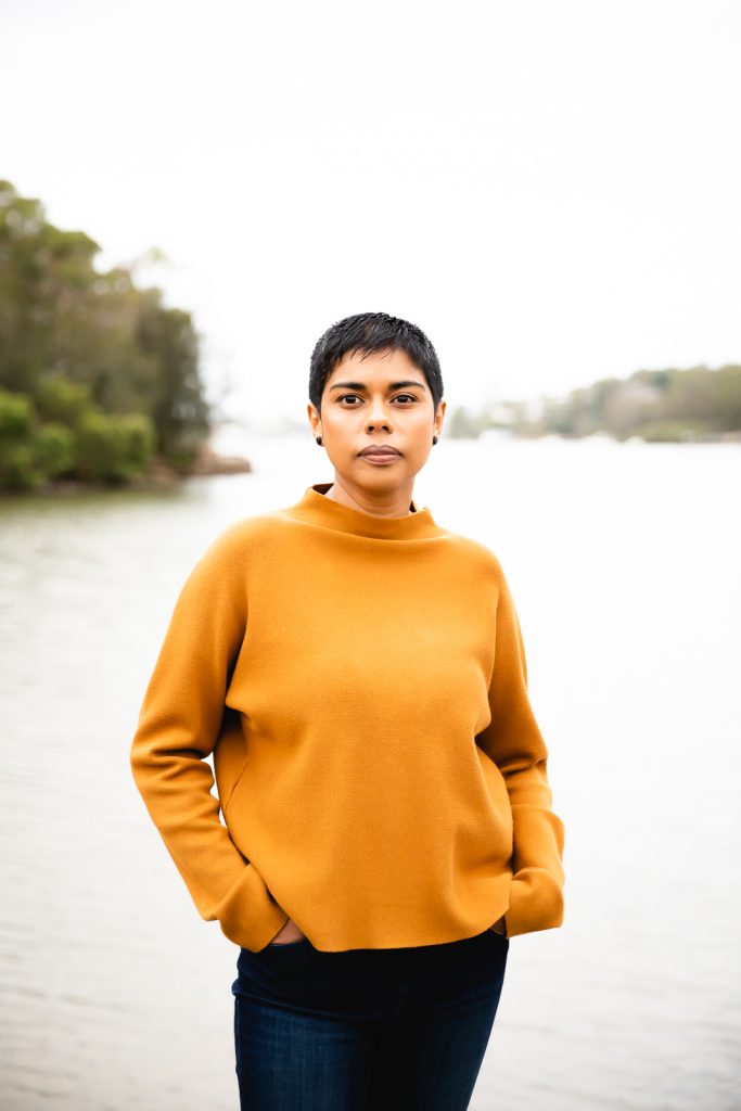 Colour publicity photo of Dinuka McKenzie standing with a river in the background. Dinuka has short black hair, brown skin and is wearing a tan long sleeved top and blue jeans.